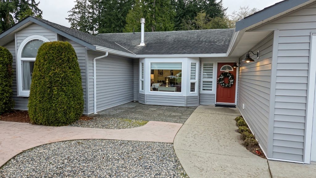 Single-storey Sechelt home re-sided in light grey vinyl with white trim around a bay window, arched window and red front door