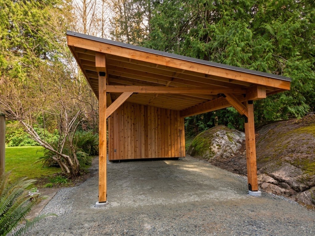 Timber-frame carport with knee-braced posts on concrete piers, open to a gravel pad, with the new cedar shed at the back